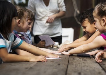 Um grupo de crianças realiza atividades educativas ao ar livre a jogar cartas alegremente em cima de uma mesa com a educadora a observar