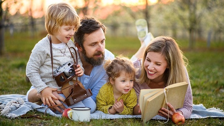 Pai, mãe e os seus dois filhos, ao ar livre e deitados numa manta, a ler alegremente um livro como atividade educativa